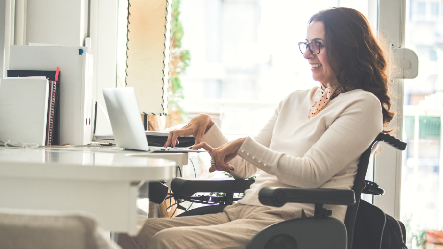 Mujer adulta con discapacidad, sentada en silla de ruedas, usa su computador portátil mientras sonríe