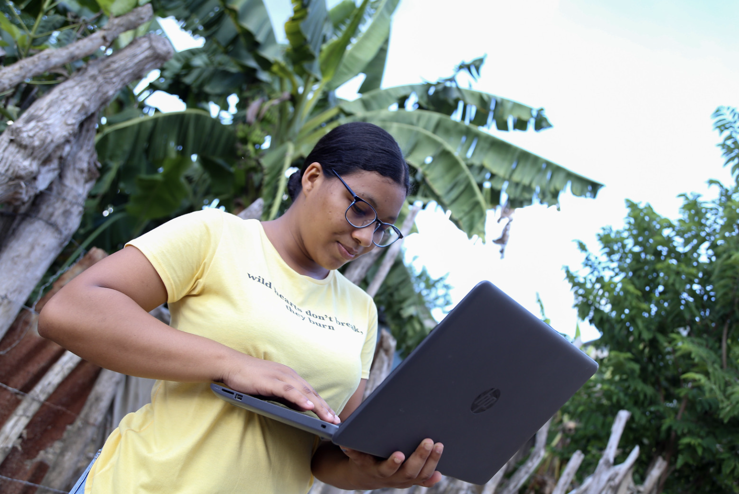 Una joven en zona rural, usa el computador portátil de pie. Con su mano izquierda lo sostiene y con la derecha, digita en el teclado.