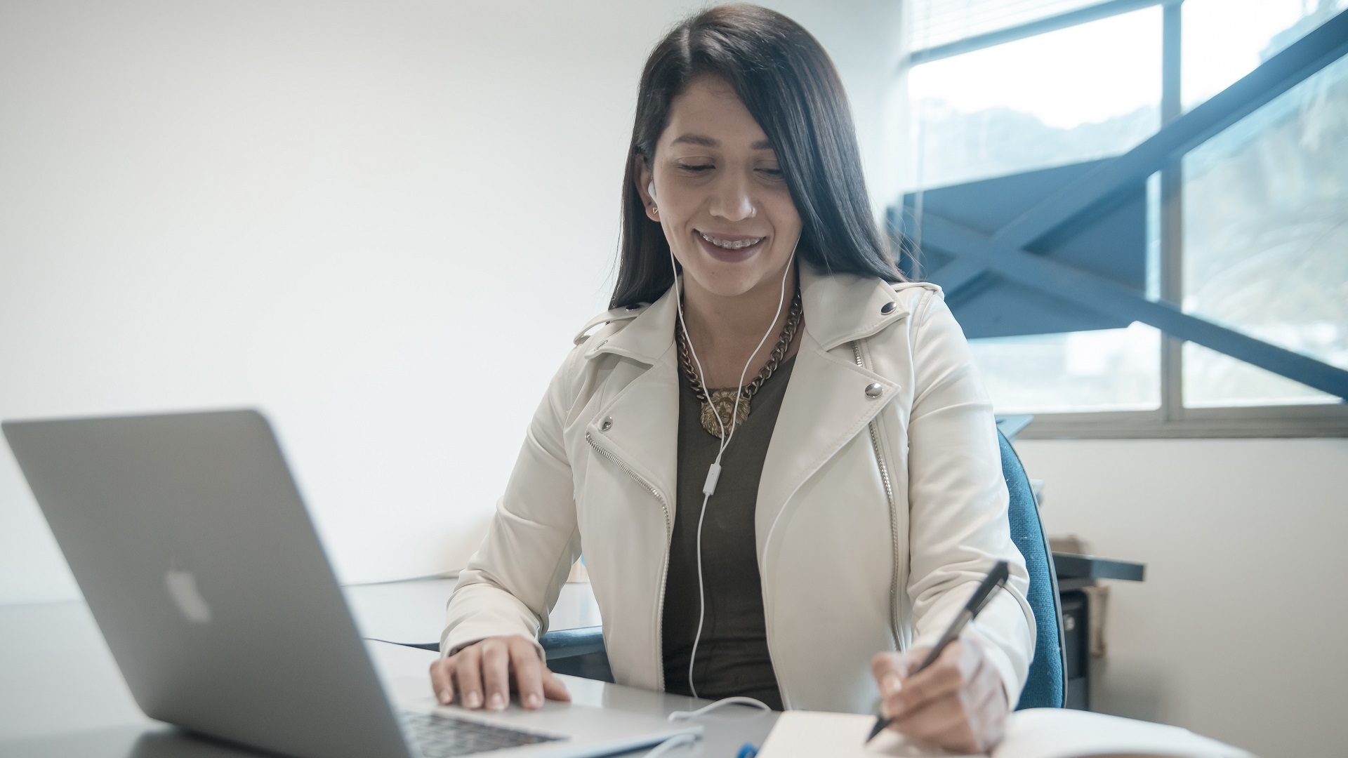  Mujer sonríe, frente a un computador, mientras escribe en una agenda lo que investigó en Internet.